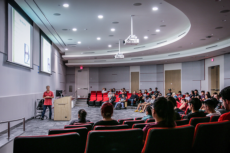 An instructor presenting at the front of an auditorium. Students fill the red auditorium seats