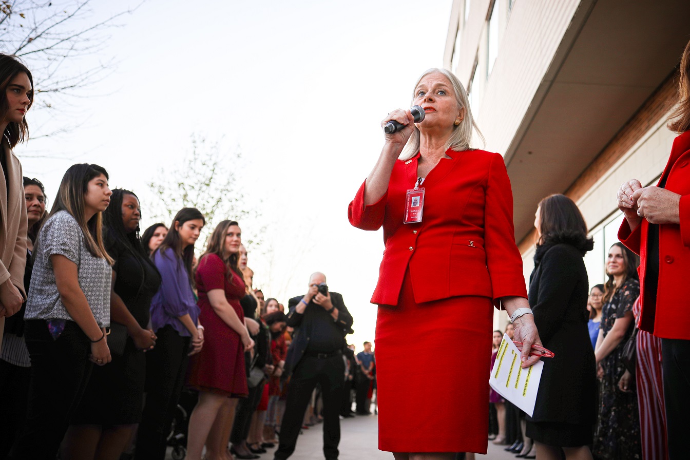 A woman in a red suit speaks into a microphone while people are a large group of people stand outside