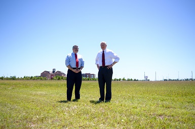 Two men in business attire standing in a grass field with the campus buildings in the background