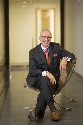 A man in glasses and a black suit smiling while sitting in a chair in hallway