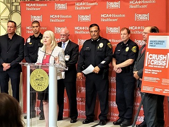 A blond woman in a suit speaks at a podium as a group of men in suits and police uniforms stand behind her
