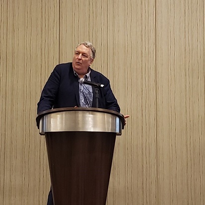 A man in a navy blue jacket standing behind a podium with a microphone.