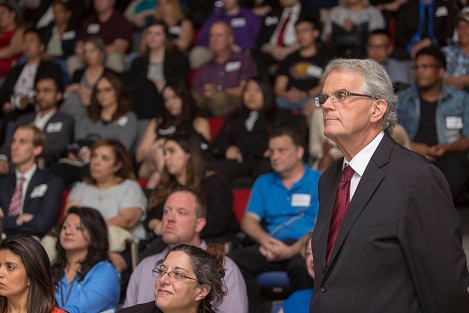 A grey-haired man wearing glass and a suit standing among people sitting in an auditorium
