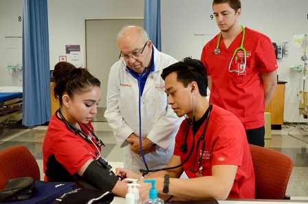 A bald male instructor wearing a white labcoat oversees a male student take a female student's blood pressue