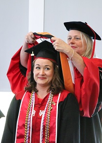 A woman in red and black academic robes places an academic hood over a woman sitting