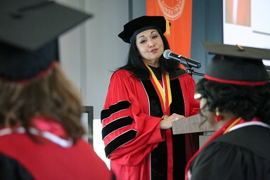 A raven-haired woman in red and black academic robes speaks at a podium with a microphone