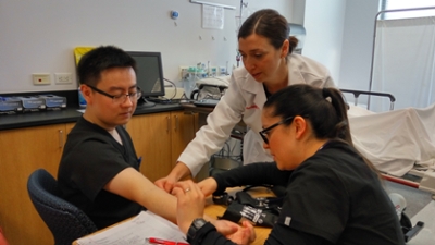 A female instructor in a labcoat oversees two students practicing with blood collection equipment