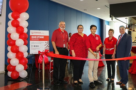 Four men and women stand behind a red ribbon with a man holding a large open pair of scissors over the ribbon