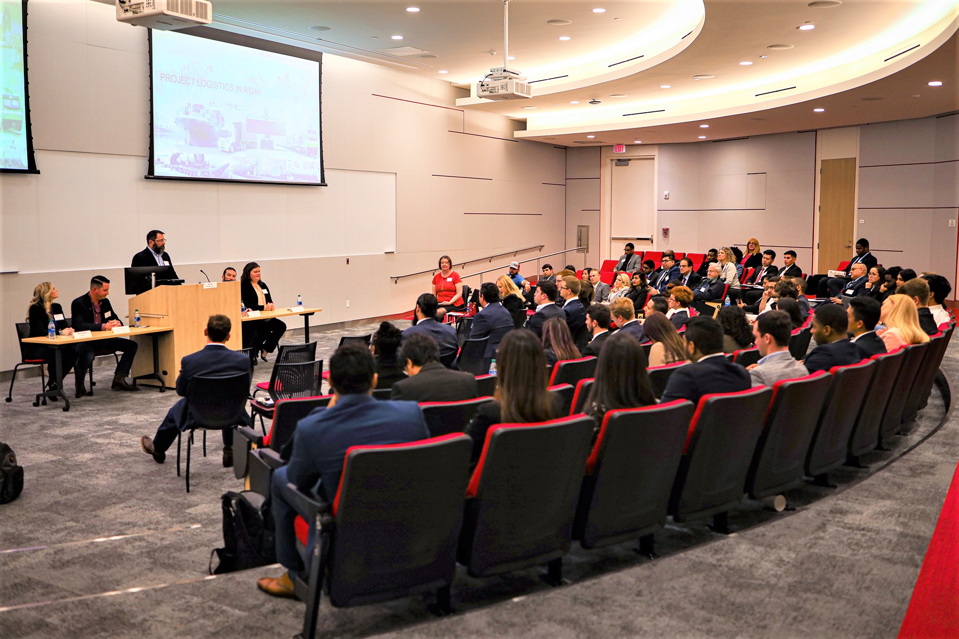 A group of people sit at the front of an auditorium classroom filled with students in business attire