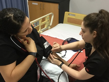 A female nursing student in black scrubs takes another female student's blood pressure