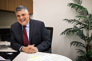 A man in dark suit smiling while sitting at a desk in an office.