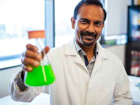 An Indian man smiling with short beard holds a flask of bright green cyanobacteria.