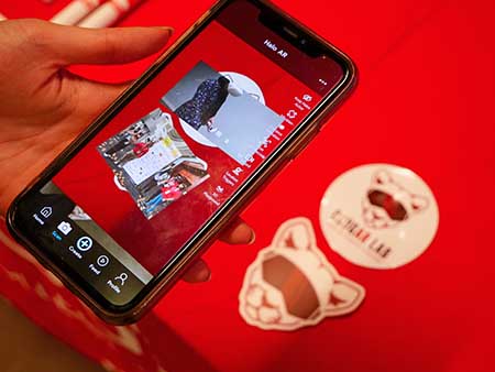 Close-up of a person holding a phone above two small printed graphics on a red table. Using its camera, the phone screen shows different images overlayed on the print graphics.