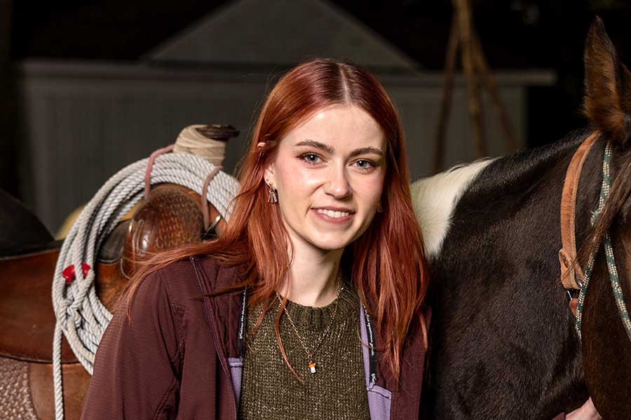 Portrait of a red-haired woman smiling and standing next to a brown horse.