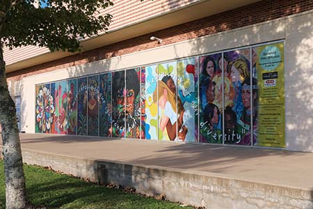 A mural of six pieces of art cover the exterior windows of a large tan brick building. A verse from a poem is superimposed over the art.