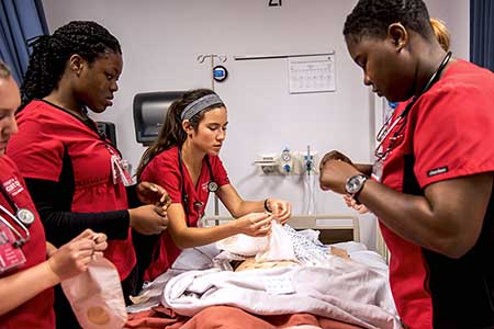 Five nursing students flank a hospital bed while preparing medical supplies.