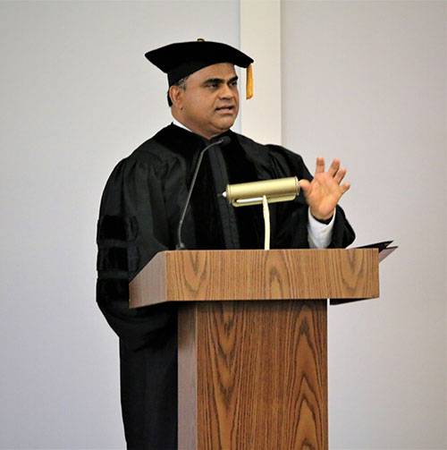 A man wearing black graduation regalia stands behind a podium.