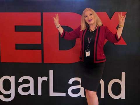 A woman in red business jacket and black dress stands in from of a large sign reading "TEDx Sugar Land"