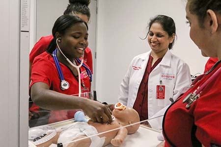 A group of nursing student wearing red scrubs inspect a baby-sized simulation mannequin. An instructor in a white lab coat oversees.