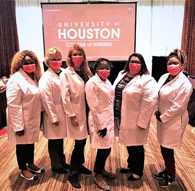 A groupd of women wearing red surgical masks and white labcoats stand in front of large screen that says, "Universiyt of Houston College of Nursing"
