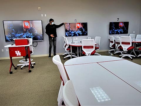 Groups of white tables with wall-mounted monitors surround a moveable podium.