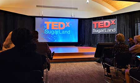 A group of people sit in darkness in front of an illuminated stage with screen displaying "TEDx Sugar Land"