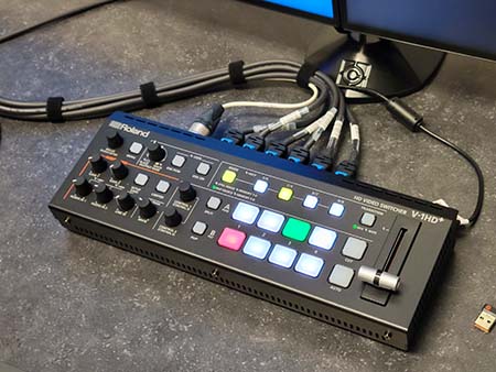 Close up of a piece of equipment used to switch video feeds to monitors. It is shaped as a rectangular prism with many knobs and illuminated push buttons.