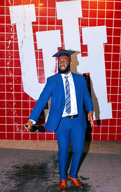 A black man wearing a blue suit and a black graduation cap opens a bottle of champagne in front of a white University of Houston logo on a red tile wall.