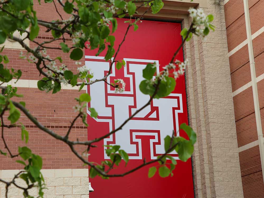 A close up of large red UH banner on the side of a brick building. Tree branches with white flowers blooming are in the foreground. .jpg