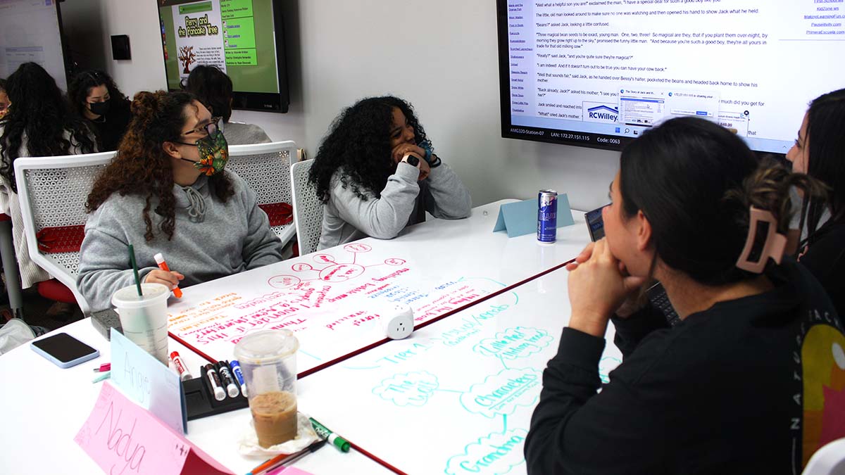 Students sitting at groups of classroom tables with a large monitor studying