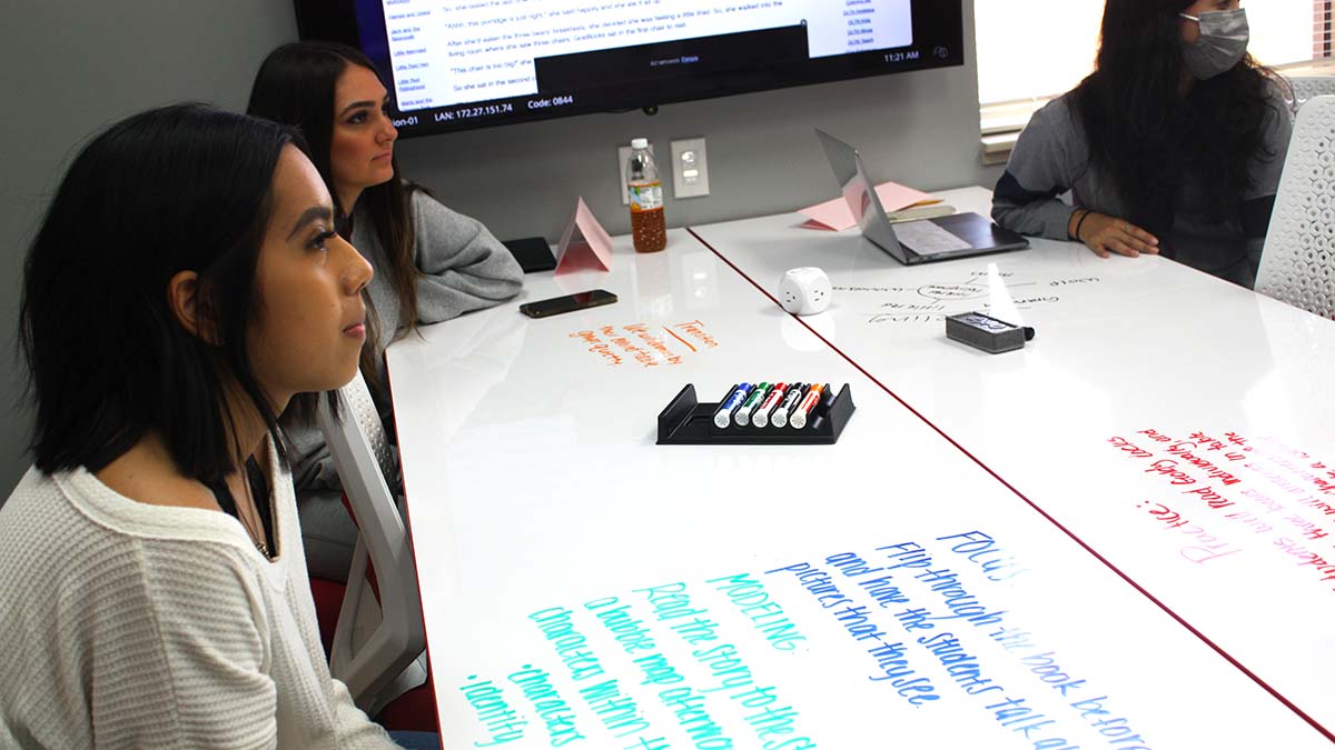 A group of students sitting at a group classroom tables with hand written notes on the whiteboard tabletop.