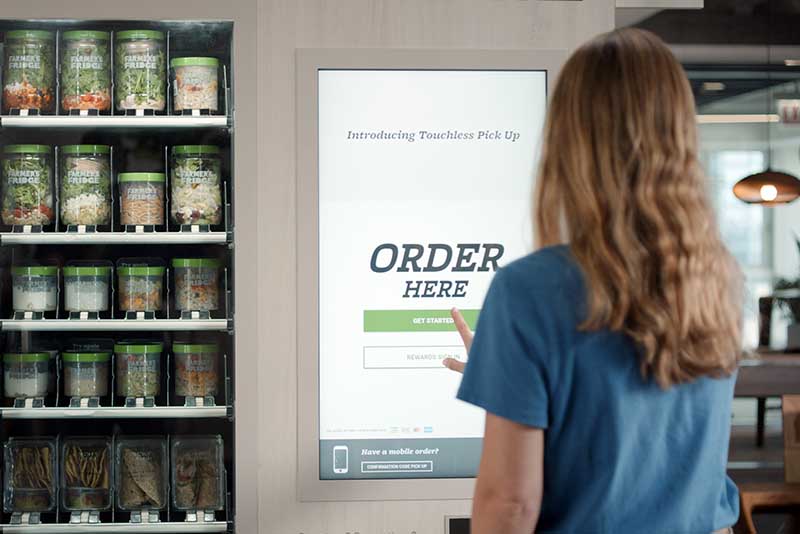 A person presses the touch screen of a vending machine. Containers of healthy food inside the machine are to the left of the screen.