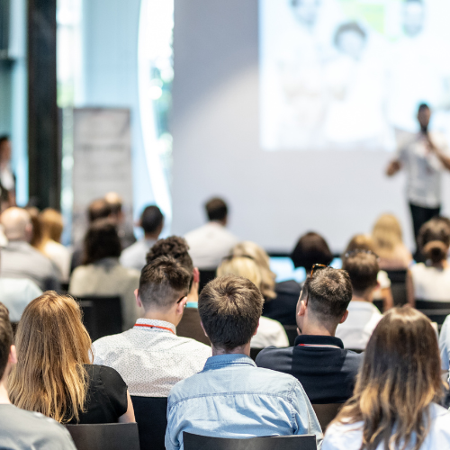 A man stands at a podium, presenting to an engaged audience seated in rows, with a projector screen in the background.